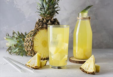 Bebida fermentada de tepache junto a pedazos de piña y una piña partida por la mitad en un fondo gris