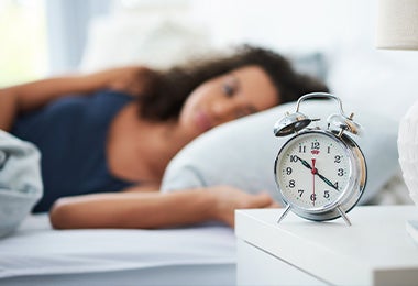 Mujer durmiendo y un reloj de alarma a su lado, lo que regula sus horas de sueño y le da un buen descanso.