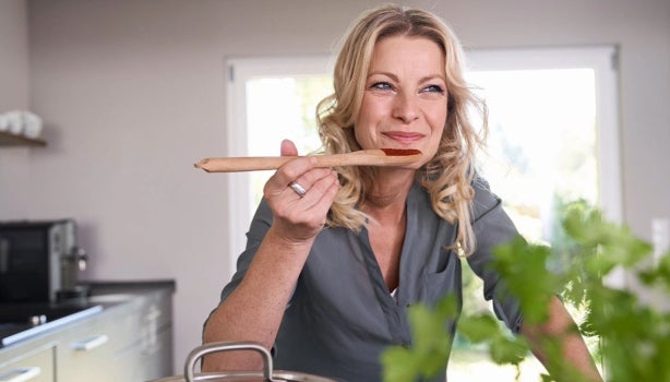 Mujer sonriente probando una cucharada de comida con salsas coreanas directamente de una olla en una cocina bien iluminada
