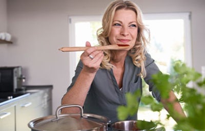 Mujer sonriente probando una cucharada de comida con salsas coreanas directamente de una olla en una cocina bien iluminada