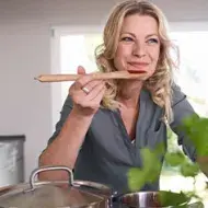 Mujer sonriente probando una cucharada de comida con salsas coreanas directamente de una olla en una cocina bien iluminada